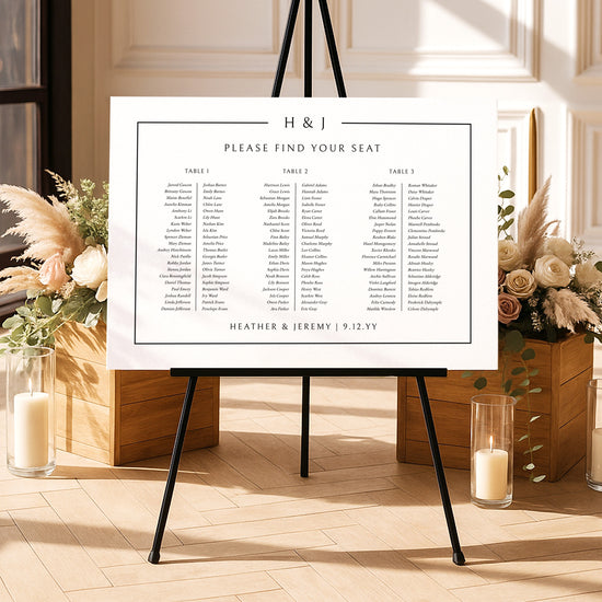 Modern long tables seating chart template on an easel with floral arrangements and candles in a decorative setting.