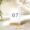 A printable table number template displayed on a small wooden block holder on a table with decorative items in the background.