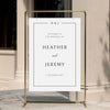 Minimal wedding welcome sign displayed on a gold stand against a neutral wall.