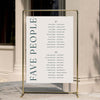 Two long tables wedding seating plan with 'FAVE PEOPLE' text on a gold stand against a neutral wall.