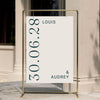 Editorial-inspired wedding welcome poster hanging from a gold pipe frame. This typographic Palais design features a chic cream and navy palette, perfect for a luxury outdoor reception. 