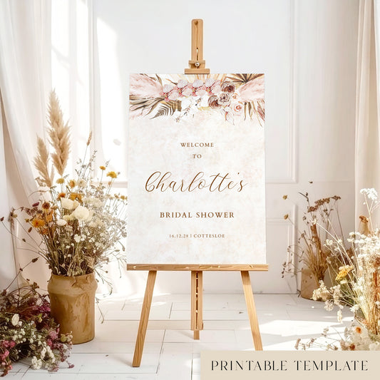 Bridal shower welcome sign with pink floral design on an easel with floral decorations and dried plants.