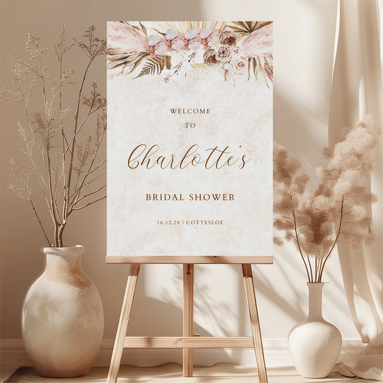 Bridal shower welcome sign with pink floral design on an easel with floral decorations and dried plants.