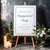 Engagement party sign on an easel with floral decorations and candles against a dark background.