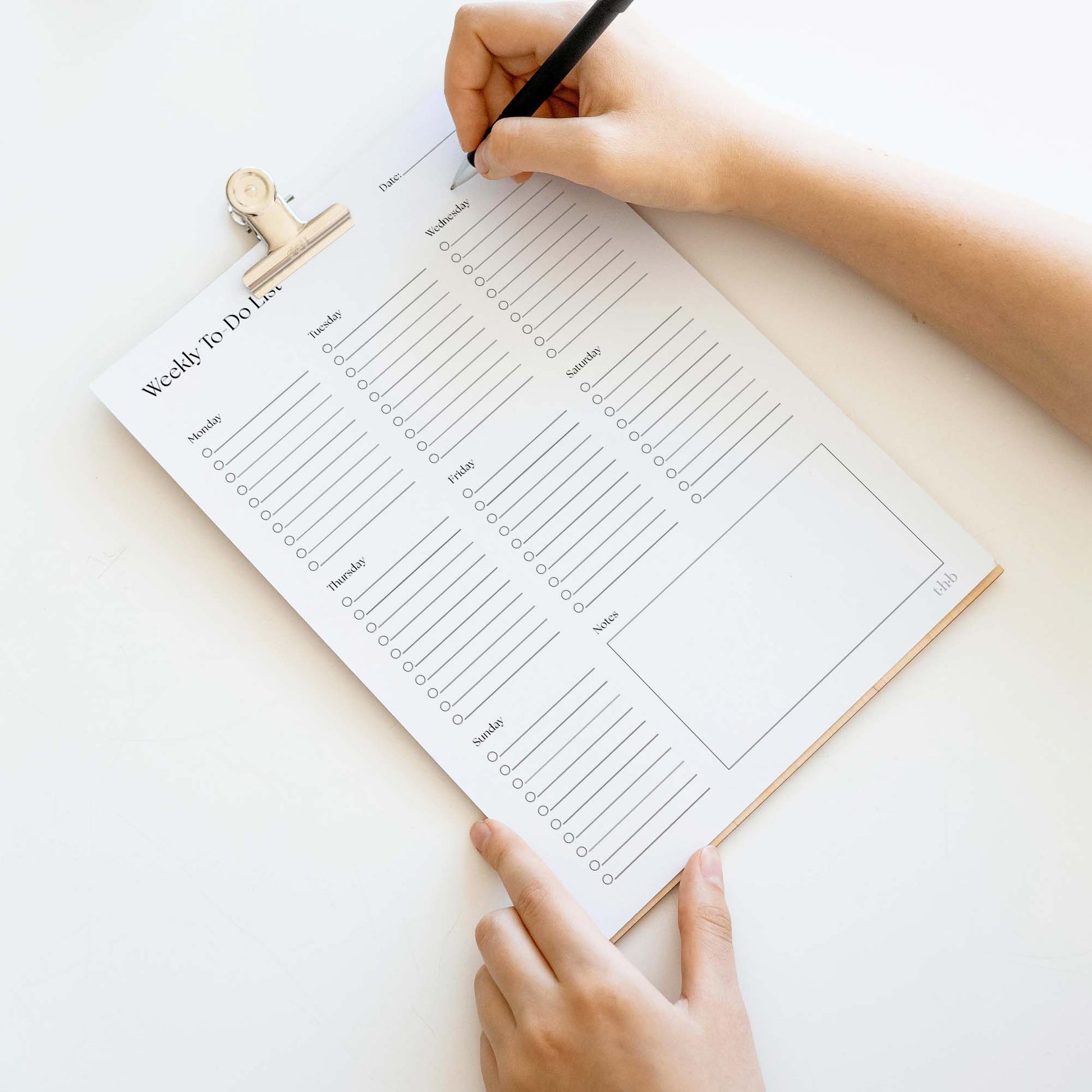 Weekly to-do list on a clipboard with hands holding a pen, on a light background.