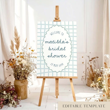 Blue whimsical bridal shower welcome sign on an easel with floral arrangements in a room setting.
