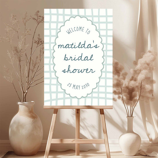 Blue whimsical bridal shower welcome sign template on an easel with floral arrangements in a room.