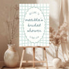 Blue whimsical bridal shower welcome sign template on an easel with floral arrangements in a room.