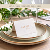 A folded place card with a name printed on it sitting on top of two plates with a eucalyptus leaf for decoration.