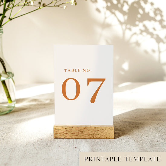 A printable table number template displayed on a small wooden block holder on a table with decorative items in the background.