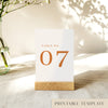 A printable table number template displayed on a small wooden block holder on a table with decorative items in the background.