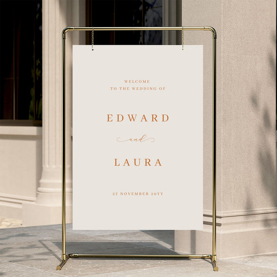 Minimal wedding welcome sign displayed on a gold stand against a neutral wall.