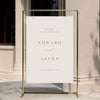 Minimal wedding welcome sign displayed on a gold stand against a neutral wall.