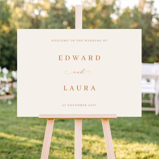 Horizontal wedding welcome sign on an easel with a blurred outdoor ceremony background.