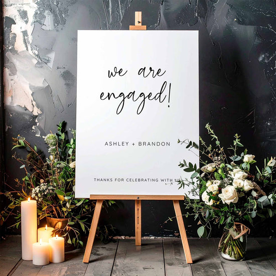 Engagement party welcome sign on an easel with floral arrangements and candles against a textured wall.