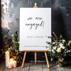 Engagement party welcome sign on an easel with floral arrangements and candles against a textured wall.