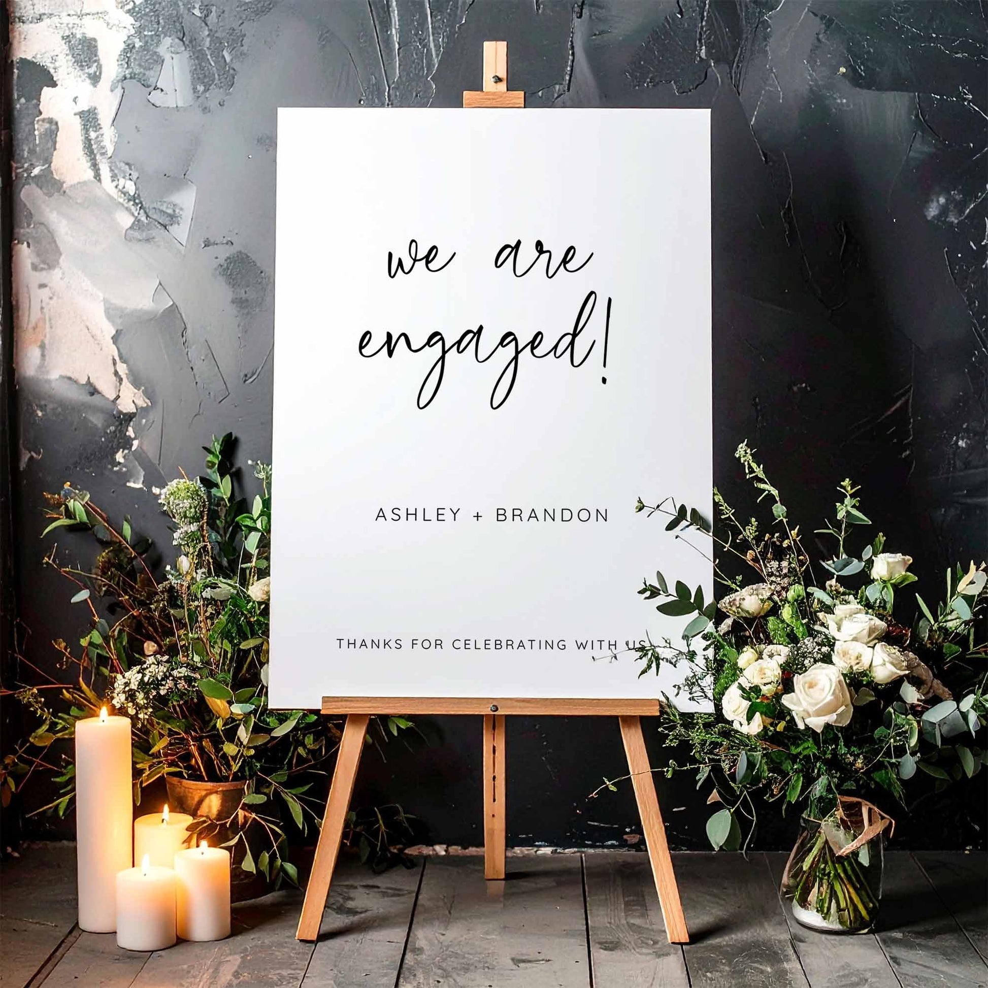 Engagement party welcome sign on an easel with floral arrangements and candles against a textured wall.