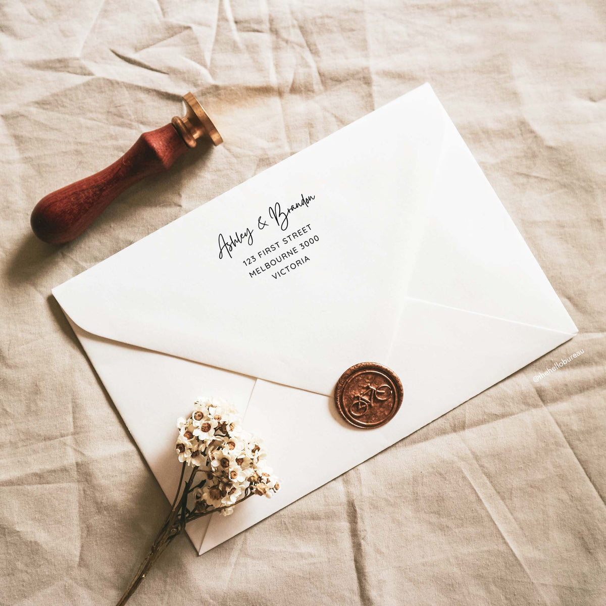 Casual handwriting wedding envelope return address template printed on the back flap of a white envelope. A copper wax seal with a bicycle, a wooden stamp and dried white flowers sit on beige linen. The minimal black text is editable in Canva.
