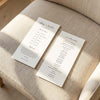Two DL-sized wedding program cards laid flat on a cushioned chair with sunlight streaming across the linen fabric.