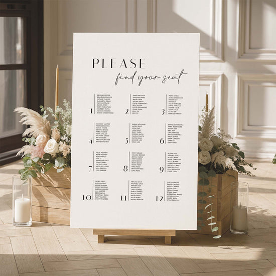 Seating chart sign with floral arrangements and candles in a room with large windows.
