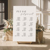 Seating chart sign with floral arrangements and candles in a room with large windows.