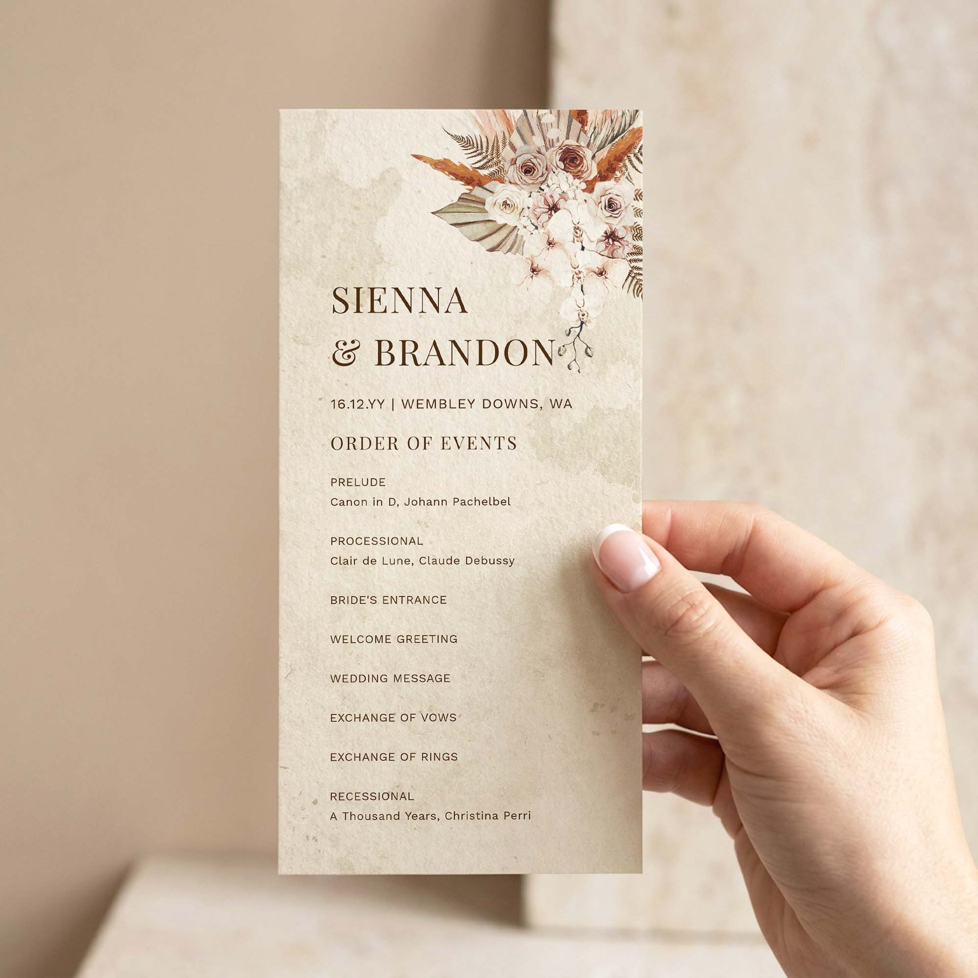 Hand holding a DIY boho wedding ceremony program template with dried floral arrangement, earthy watercolour texture and order of events listing.