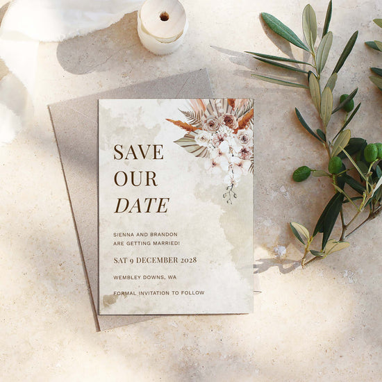 Modern boho save the date template design printed on a white card sitting on a textured surface with olive branches.