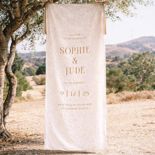 Decorative wedding fabric welcome sign hanging from a tree in a scenic outdoor setting.
