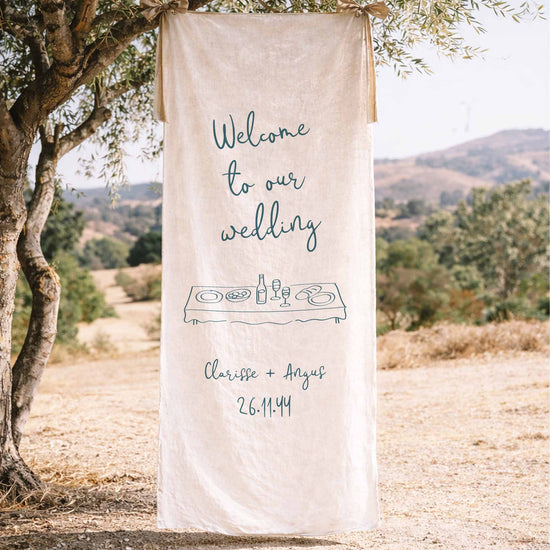 Decorative illustrated wedding fabric welcome sign hanging from a tree in a scenic outdoor setting.