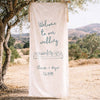 Decorative illustrated wedding fabric welcome sign hanging from a tree in a scenic outdoor setting.